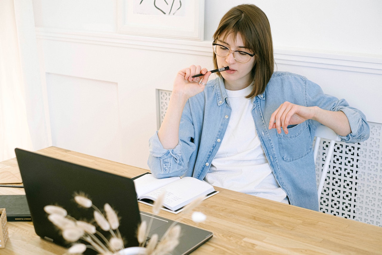 Lady with pen near mouth and open notebook in front of laptop